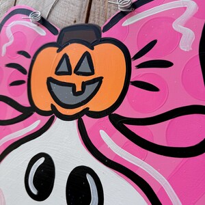 May include: A pink and white ghost with a black outline and a pumpkin on its head. The pumpkin has a black outline and a smiling face with triangle eyes. The ghost has black eyes and a black mouth.