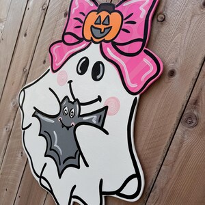 May include: A white ghost with a pink bow and a pumpkin on top, holding a gray bat in its arms. The ghost has a smiling face with two black eyes and a black mouth.