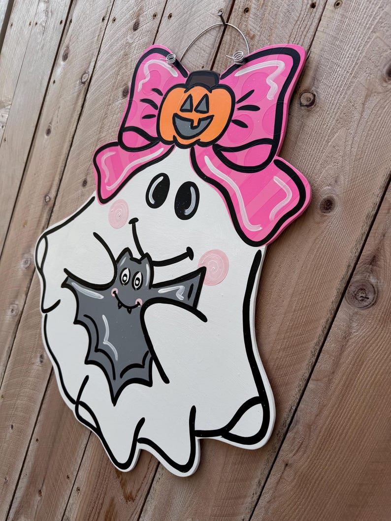 May include: A white ghost with a pink bow and a pumpkin on top, holding a gray bat in its arms. The ghost has a smiling face with two black eyes and a black mouth.