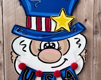 May include: A wooden door hanger shaped like a cartoon Uncle Sam with a blue top hat, red and white stripes, and a yellow star. The hanger has a red, white, and blue banner with the word "USA" on it.