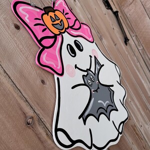 May include: A white ghost with a pink bow and a black bat in its arms. The ghost has a smiling face with two black eyes and a pink nose. The bat has a smiling face with two black eyes and a pink nose. The ghost is holding the bat in its arms and the bat is looking up at the ghost.
