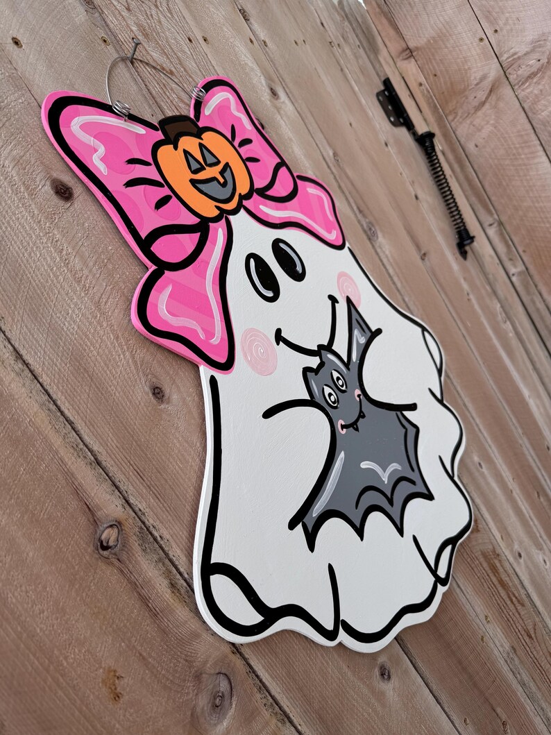 May include: A white ghost with a pink bow and a black bat in its arms. The ghost has a smiling face with two black eyes and a pink nose. The bat has a smiling face with two black eyes and a pink nose. The ghost is holding the bat in its arms and the bat is looking up at the ghost.