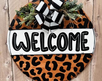 May include: Round wooden door hanger with a black and white striped bow and greenery on top. The bottom half of the door hanger is painted with a brown and black leopard print. The top half is white with the word "WELCOME" written in black.