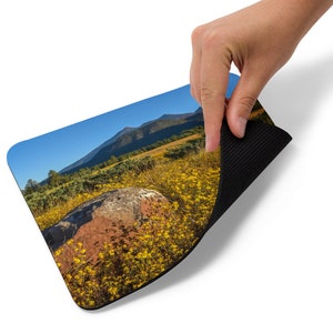 May include: A mouse mat featuring a scenic image of a large rock in a field of yellow wildflowers with a mountain range in the background. The mouse mat has a black rubber backing.