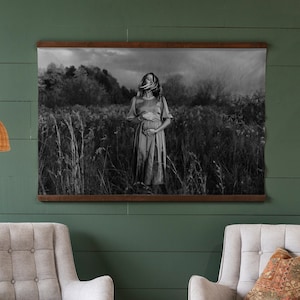 May include: A black and white photograph of a woman standing in a field of tall grass. She is wearing a long dress and has her hands on her stomach. The photo is framed in a dark wood frame and hanging on a green wall.