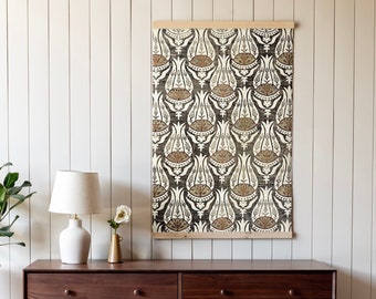 May include: A dark brown wooden dresser with six drawers and brass knobs. The dresser is sitting on a light wood floor in front of a white wall with a patterned tapestry hanging above it. A lamp and a small vase with a white flower are on the top of the dresser.