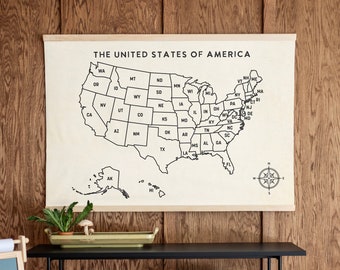 May include: A black metal console table with two shelves. The top shelf has a green tray and a small potted plant. The bottom shelf has a woven basket, two brown storage boxes, and a brown fedora hat. A black and white map of the United States hangs on the wall behind the table. The map is labeled "The United States of America".
