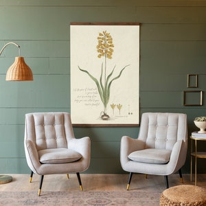 May include: A botanical print with a yellow flower and green leaves, framed in a wooden frame, hangs on a green wall. The print includes a bible verse: "Let the peace of Christ rule in your hearts, to which also you were called in one body; and be thankful."  Two gray armchairs with tufted upholstery and gold legs are positioned in front of the print. A small gold side table with a book and a white ceramic bowl with green plants sits next to one of the chairs. A woven brown ottoman sits in front of the chairs.
