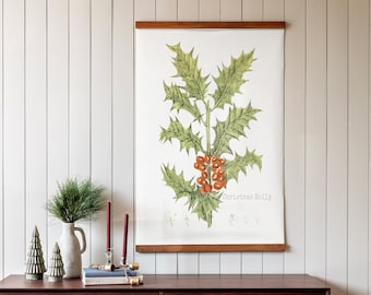 May include: A framed botanical illustration of a Christmas holly plant with red berries. The illustration is on a white background and has the text "Christmas Holly" printed below the image. The frame is brown wood and the illustration is hanging on a white wall above a dark brown dresser with drawers.
