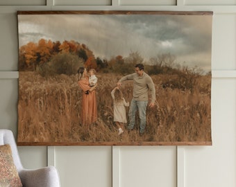 May include: A family of four pose in a field of long grass. The parents are holding their two children, one in each arm. The parents are smiling and looking at each other. The children are looking at the camera. The background is a field of long grass and trees. The sky is cloudy.