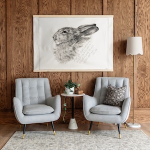 May include: Two gray armchairs with black legs and a small round side table with a black base and a white top. The chairs are upholstered in a light gray fabric and have a tufted design. The table is decorated with a small potted plant and a book. A framed print of a rabbit with a quote about the rabbit is hanging on the wall behind the chairs.