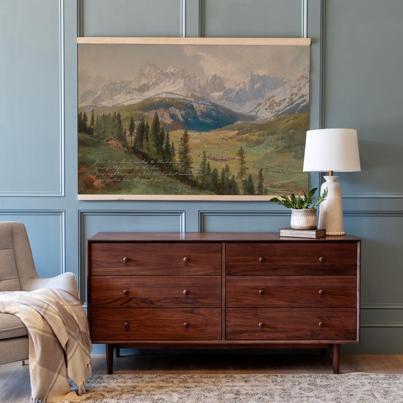 May include: A landscape painting of a mountain range with text at the bottom is displayed above a dark wood dresser. A white lamp and a small potted plant sit on the dresser. A gray armchair and a patterned rug are in the foreground.
