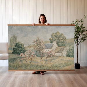 May include: A large landscape painting with a wooden frame. The artwork depicts a rural scene with a house, trees, and a field. The color palette is soft, with muted greens, blues, and pinks. The painting is held up by a person.