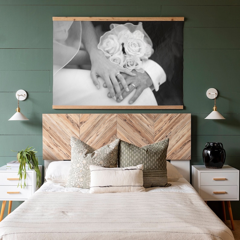 May include: A black and white photograph of a couple holding hands with a bouquet of flowers. The photo is framed and hanging above a wooden headboard with a chevron pattern. The headboard is on a bed with white bedding and two throw pillows.