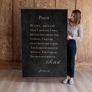 May include: A black wall hanging with the text of Psalm 46:10-11 in white script. The wooden frame is a medium brown. The text includes the words "Be still, and know that I am God".