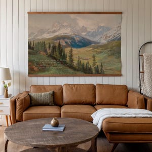 May include: A large landscape tapestry depicting a mountain range with snow-capped peaks, a valley, and evergreen trees. The tapestry is mounted on a wooden rod and hangs above a brown leather sectional sofa. The text is visible on the tapestry.
