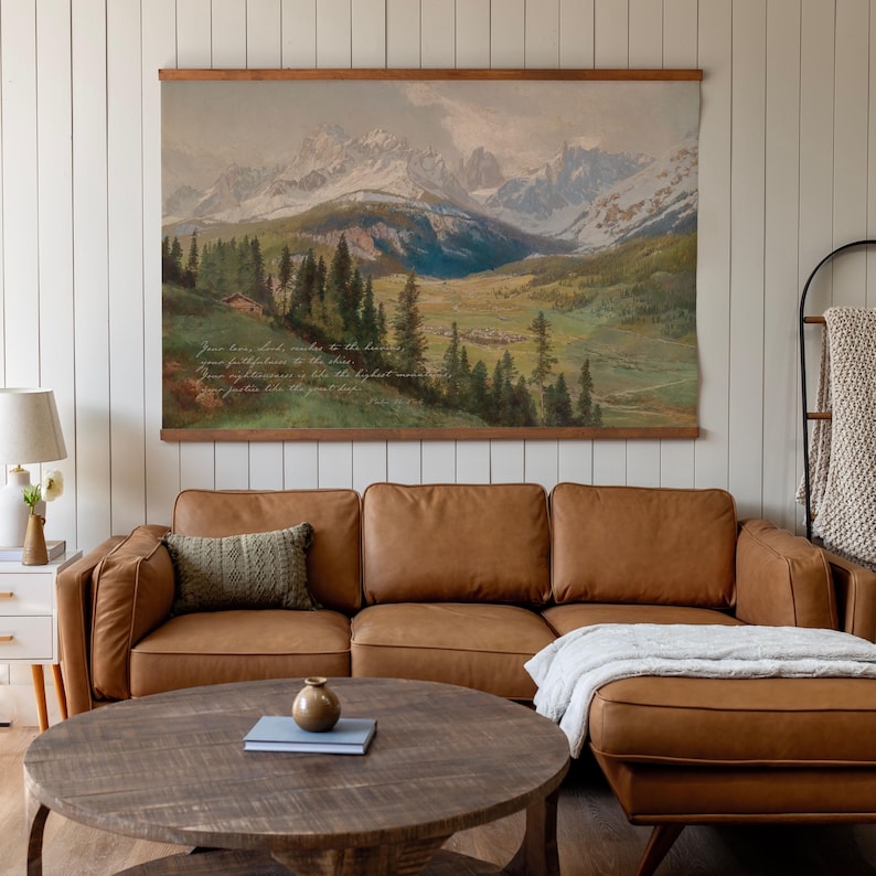May include: A large landscape tapestry depicting a mountain range with snow-capped peaks, a valley, and evergreen trees. The tapestry is mounted on a wooden rod and hangs above a brown leather sectional sofa. The text is visible on the tapestry.