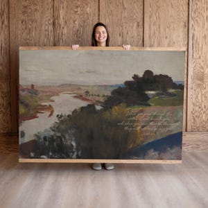 May include: A framed landscape painting with a river and trees, in shades of brown, green, and gray. The artwork has a light-colored wooden frame. Text on the painting reads, "When you pass through the waters, I will be with you..."