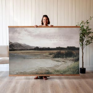 May include: A large landscape print with a wooden frame. The artwork depicts a serene scene with a lake, mountains, and a cloudy sky. The print includes a quote in cursive script. The overall color palette is muted with shades of gray, brown, and green.