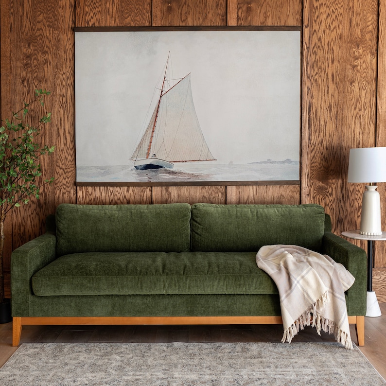 May include: A green velvet sofa with a white and beige throw blanket in front of a wall with a framed watercolor painting of a sailboat.