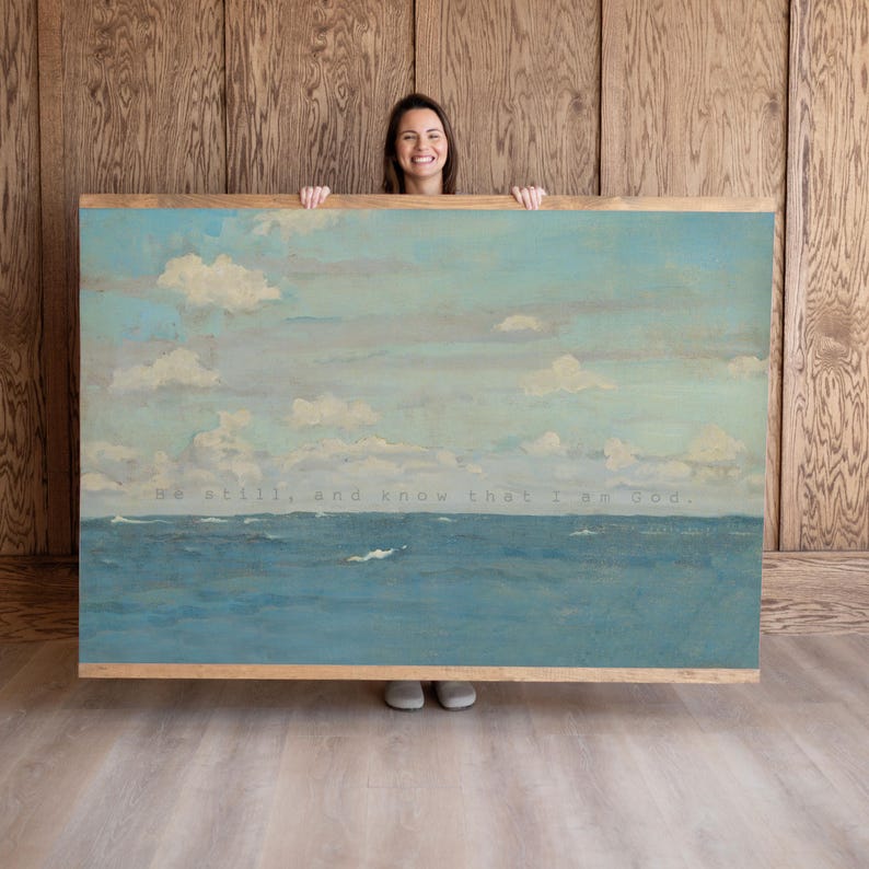 Pu&ograve; includere: Dipinto di paesaggio incorniciato raffigurante una serena scena oceanica sotto un cielo nuvoloso. L'opera presenta sfumature di blu e bianco, con il testo "Be still, and know that I am God." La cornice &egrave; in legno chiaro.