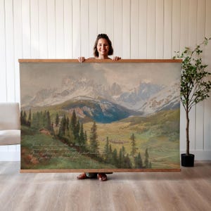 May include: A large landscape painting of a mountain range with snow-capped peaks, a valley, and a forest. The artwork is framed in a wooden border and features text in the lower left corner. The painting is held by a person.