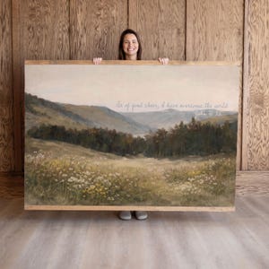 May include: A framed landscape painting with a serene meadow and mountain scene. The artwork features a quote that reads, "Be of good cheer, I have overcome the world." The painting is held by a person.
