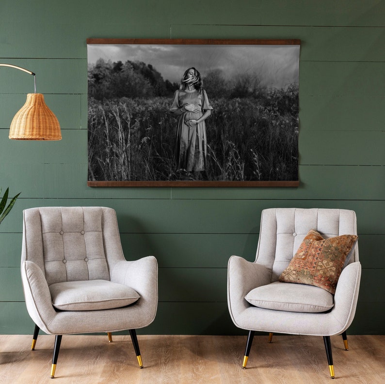 May include: A black and white photograph of a woman standing in a field of tall grass. She is wearing a long dress and has her hands on her stomach. The photo is framed in a dark wood frame and hanging on a green wall.