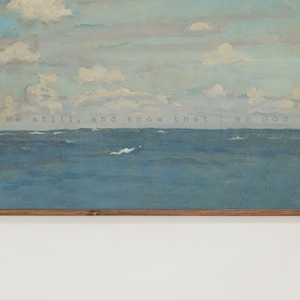 Pu&ograve; includere: Un'opera d'arte incorniciata raffigurante un sereno paesaggio marino con un cielo nuvoloso. L'oceano &egrave; dipinto in tonalit&agrave; di blu, con nuvole bianche e il testo "Be still, and know that I am God." L'opera &egrave; incorniciata in una cornice di legno marrone.