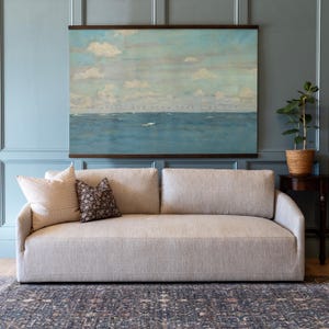 Pu&ograve; includere: Un divano in tessuto beige con due cuscini decorativi, uno a righe e uno fantasia. Sopra il divano &egrave; appeso un dipinto di paesaggio marino incorniciato con il testo "Be still, and know that I am God." Una pianta in vaso &egrave; su un tavolino.