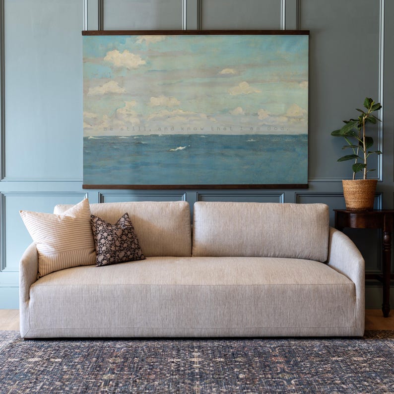 Pu&ograve; includere: Un divano in tessuto beige con due cuscini decorativi, uno a righe e uno fantasia. Sopra il divano &egrave; appeso un dipinto di paesaggio marino incorniciato con il testo "Be still, and know that I am God." Una pianta in vaso &egrave; su un tavolino.