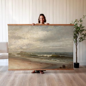 May include: A framed painting depicting a seascape with a sandy beach, foamy waves, and a cloudy sky. The painting is in a wooden frame and is hanging on a white wall.