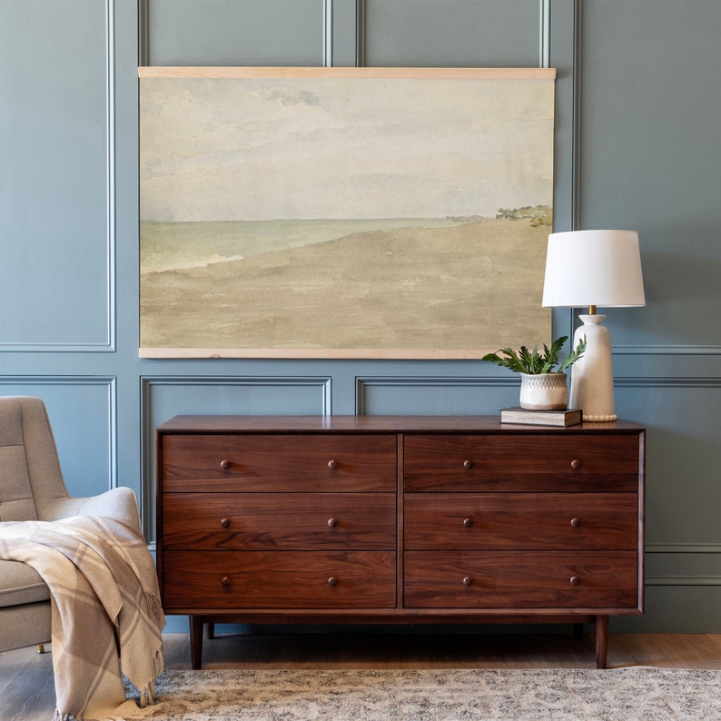 May include: A brown wooden dresser with six drawers and brass knobs. The dresser is in front of a light blue wall with a framed watercolour painting of a beach scene above it. A white lamp with a white shade is on the dresser next to a small potted plant.