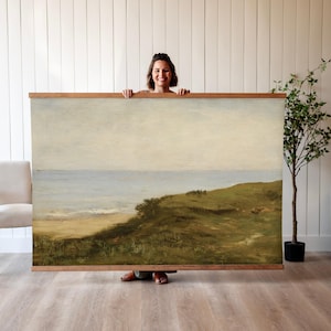 May include: A vintage landscape painting depicting a grassy hillside overlooking a calm ocean with a sandy beach. The sky is a pale blue with wispy clouds. The painting is framed in a simple wooden frame.