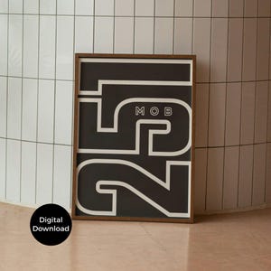 May include: A framed art print with a dark background and bold white graphic design. The design features the number "25" and the letters "MOB". The frame is a warm brown color. A black circle with the words "Digital Download" is in the lower left corner.