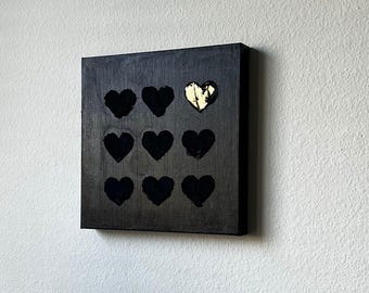 May include: Square black canvas art featuring nine heart shapes. Eight hearts are solid black, while one heart is gold. The artwork is mounted on a white wall, creating a striking contrast.