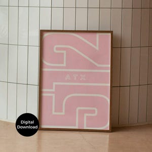 May include: A framed art print featuring the number "512" in a bold, stylized design with a light pink background and white outlines. The letters "ATX" are centered within the design. The frame is a dark brown color. A digital download is available.