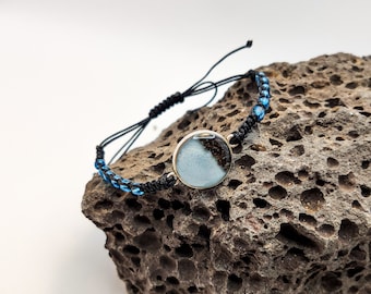 Pode incluir: Um bracelete de cordão preto com contas de vidro azul e um amuleto redondo de resina com um design branco e castanho.