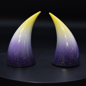 May include: Two gradient-colored horns. The tips are yellow, transitioning to white, then purple at the base. The horns are slightly curved and have a glittery finish. They are displayed on a black surface.