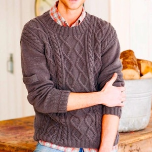 May include: A gray cable-knit sweater with a crew neck. The sweater has a textured pattern and is worn over a plaid shirt and blue jeans. The person is smiling and leaning on a wooden surface.
