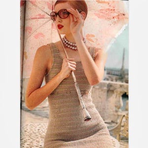 May include: A beige sleeveless crochet dress with a fitted bodice and a thin waist belt. The garment is paired with a multi-strand pearl necklace, dark-tinted sunglasses, and a large, light-colored parasol featuring a subtle pink floral pattern.