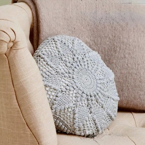 May include: A light beige upholstered armchair with a round, gray crocheted pillow and a matching throw. The pillow has a detailed, circular pattern. The armrest is curved and the chair has a button-tufted seat.