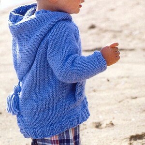 May include: A blue knitted hooded sweater with a tassel detail. The sweater has a hood, pockets, and a ribbed hem. The child is wearing a plaid patterned bottom. The sweater is made of a soft, chunky knit.