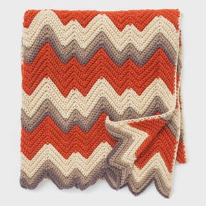 May include: A folded, handmade crochet blanket with a chevron pattern. The blanket features alternating bands of burnt orange, beige, and gray. The blanket is made of a soft, textured yarn and has a scalloped edge.