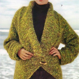 Puede incluir: Una mujer con un abrigo de punto verde y marrón con cuello chal y cierre de palanca de madera. El abrigo es de punto grueso con un aspecto texturizado. La mujer lleva un cuello alto negro.