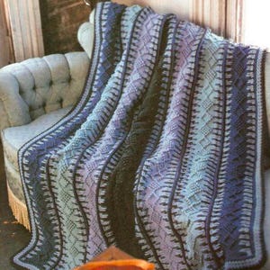 Peut inclure: Une couverture tricotée avec un motif géométrique dans les tons de bleu, de violet et de vert, drapée sur un fauteuil de couleur claire. La couverture a une bordure foncée. La texture et le dégradé de couleurs créent une esthétique chaleureuse et accueillante.