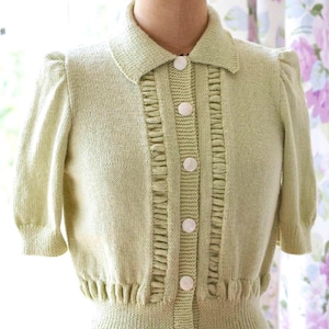 Könnte beinhalten: Hellgrüne Strickjacke mit kurzen Ärmeln, Kragen und dekorativen Rüschen auf der Vorderseite. Die Strickjacke hat große, runde, weiße Knöpfe und einen gerippten Saum. Das Kleidungsstück wird auf einer Schaufensterpuppe präsentiert.
