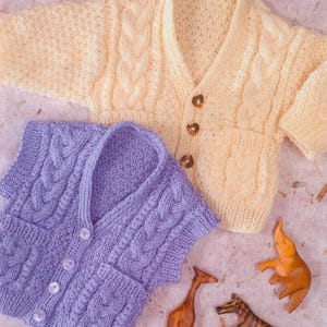 Puede incluir: Un cárdigan de punto amarillo pálido de manga larga con patrón de trenzas y tres botones marrones, y un chaleco de punto morado sin mangas con patrón de trenzas y cuatro botones blancos, junto a tres pequeñas figuras de animales de madera.
