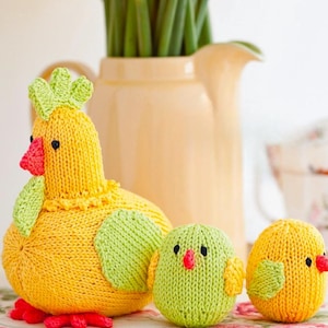 May include: A knitted yellow hen with green detailing and red feet, alongside two knitted chicks. One chick is green, the other yellow. The hen has a green crown and red beak. A vase of yellow flowers is in the background.
