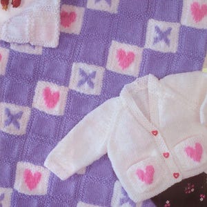 May include: A knitted baby blanket featuring a purple and white checkered pattern with alternating pink hearts and purple X symbols, paired with a white long-sleeved cardigan that has two front pockets with pink hearts and three small pink heart buttons.
